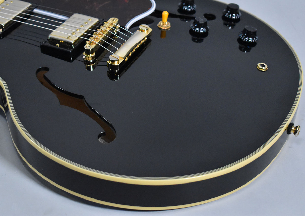 Close-up of the glossy black body, showing gold hardware, f-hole, cream binding, four black knobs, and a yellow-tipped toggle switch.
