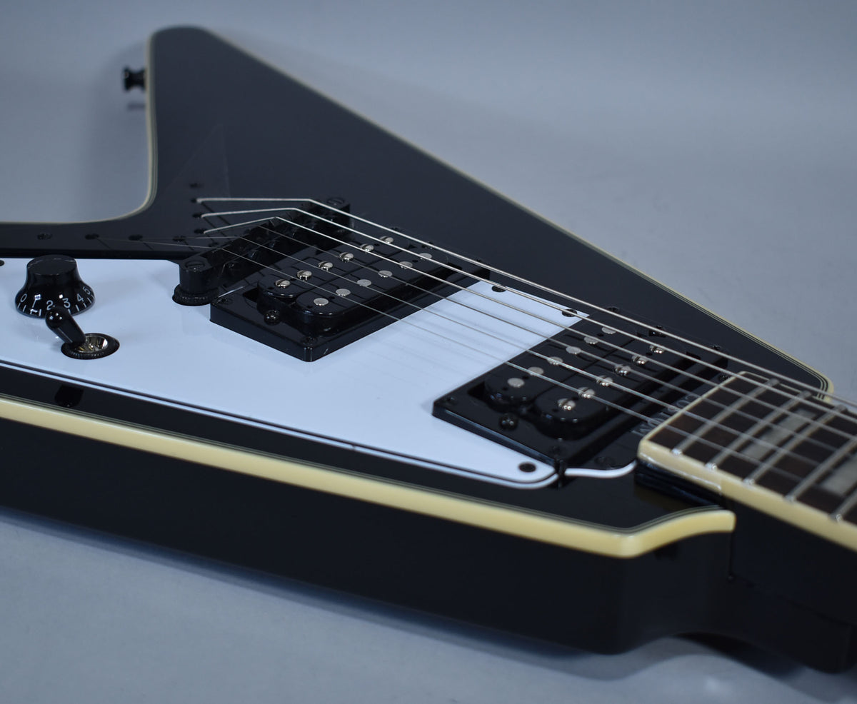 Close-up of a black Flying V guitar body, showing the white pickguard, two black humbuckers, a black knob, and a three-way switch.