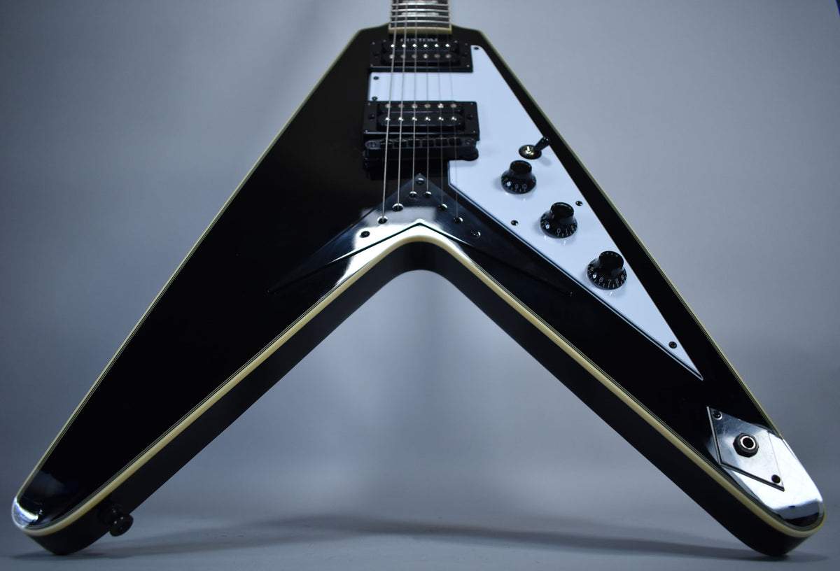 The black Flying V guitar, with a white pickguard and three black control knobs, is angled to show its body and lower neck.
