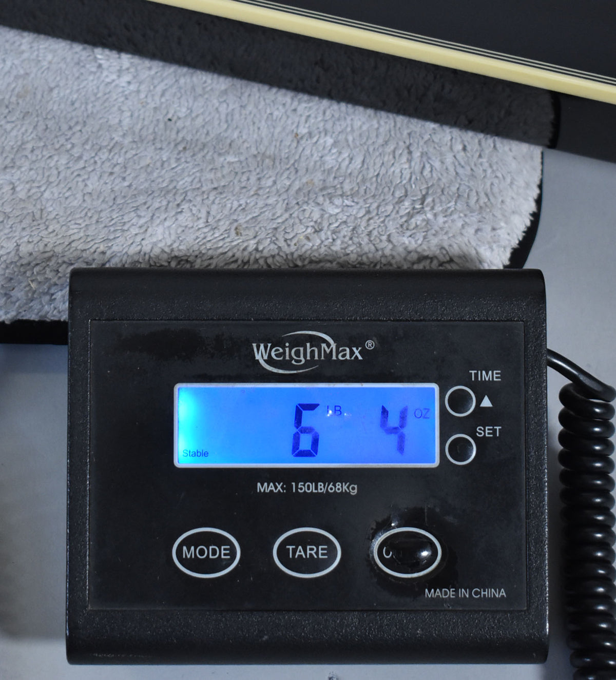 A black digital scale displays "6 LB. 4 OZ." with a guitar's black body and cream binding resting on a grey towel above it.