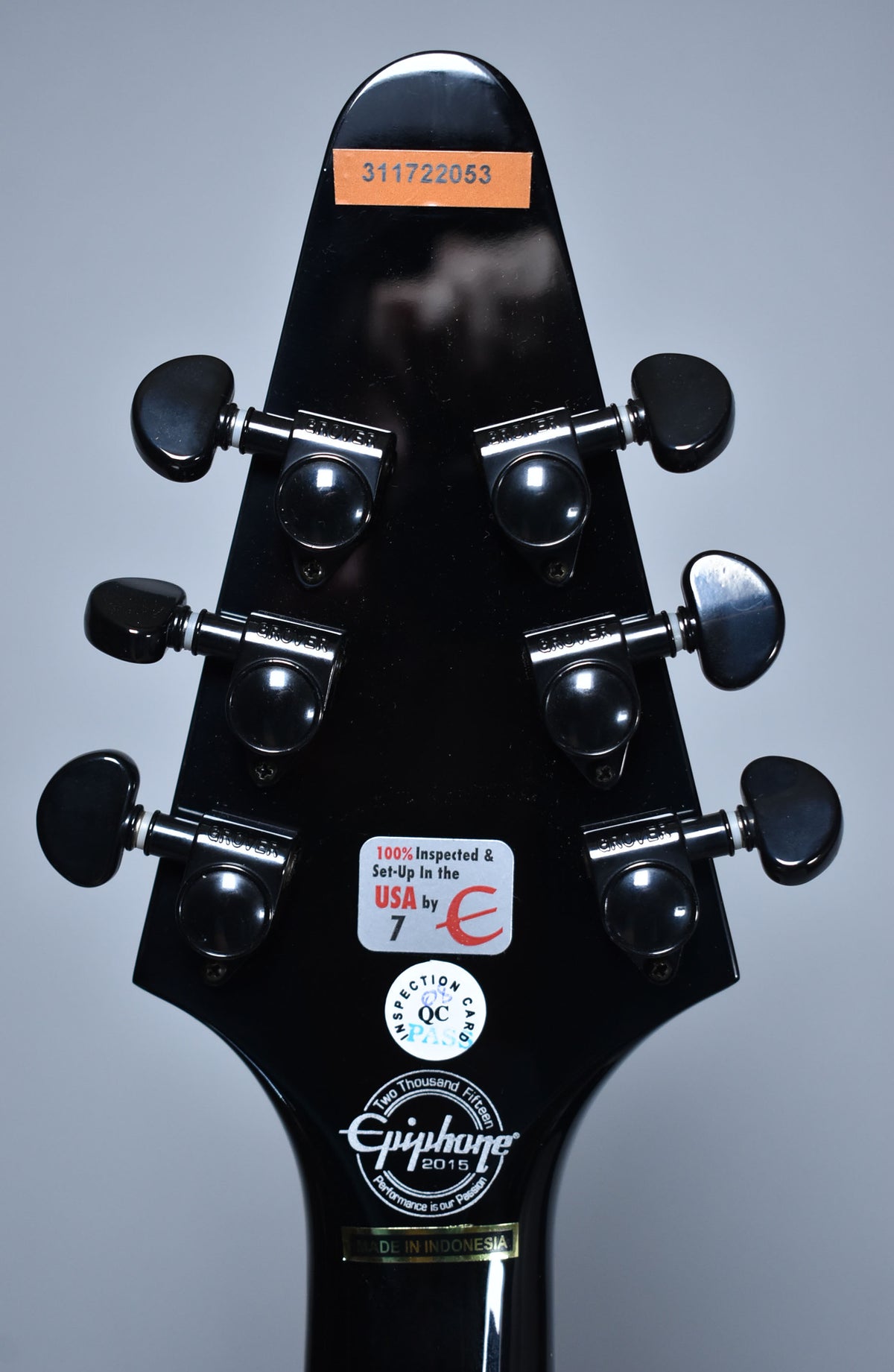 The back of a glossy black headstock with six black Grover tuners, showing various inspection stickers and a serial number.