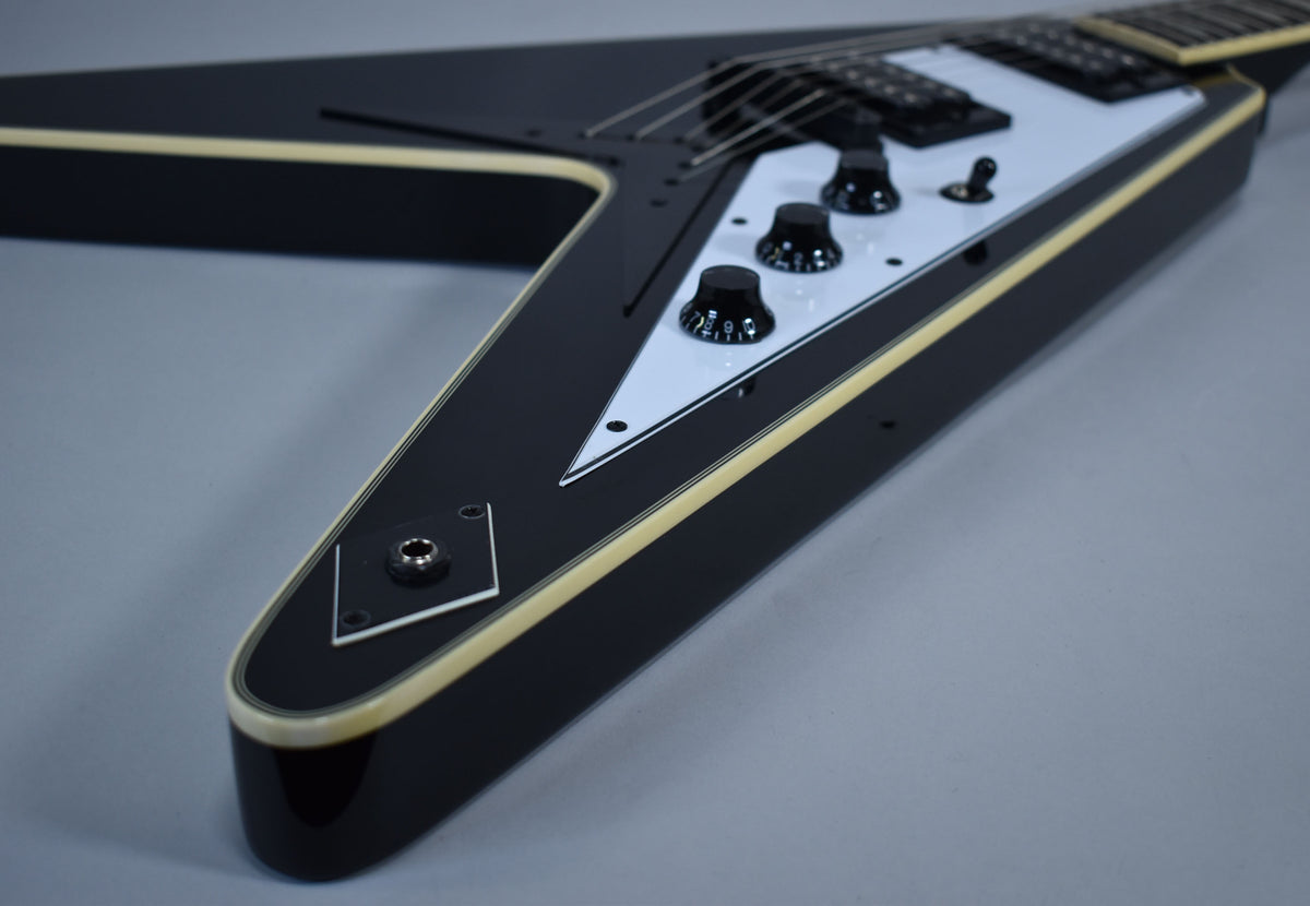 The lower wing of a glossy black Flying V, showing the white pickguard, three black control knobs, and output jack.