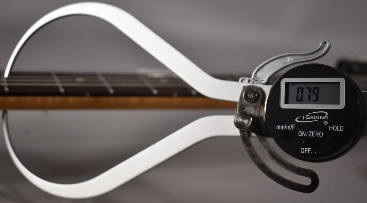 A digital caliper measures the string height of the guitar's dark fretboard and light brown neck, displaying "0.79" on its screen.