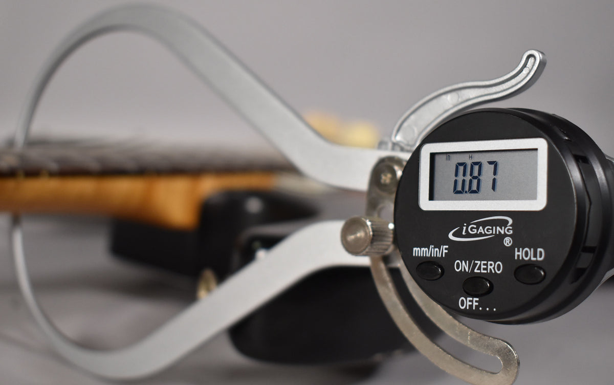 A digital caliper measures the guitar's neck relief at 0.87, with the fretboard and sunburst body blurred in the background.