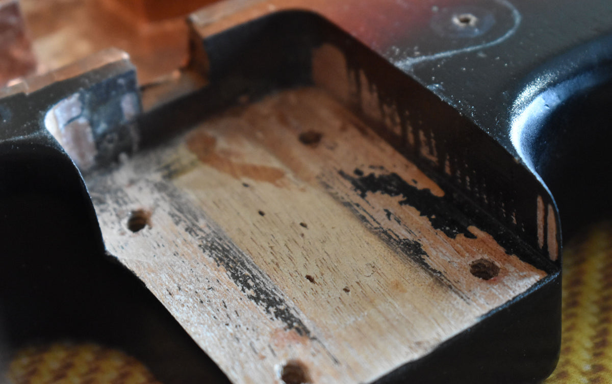 Close-up of the guitar's body cavity, showing exposed wood with black paint residue and three screw holes, surrounded by a sunburst finish.