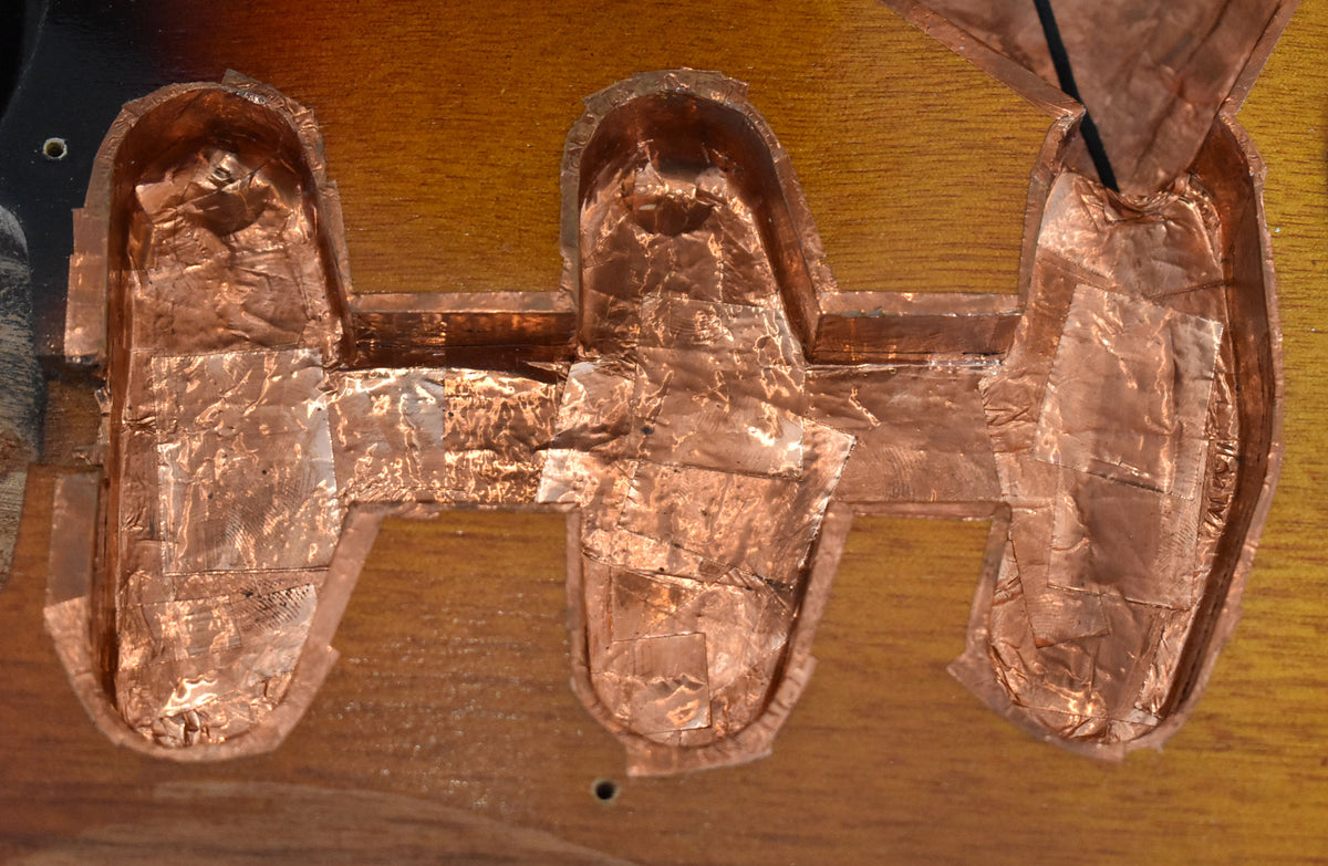 Close-up of the guitar's body, showing three pickup cavities lined with crinkled copper shielding tape on a sunburst finish.