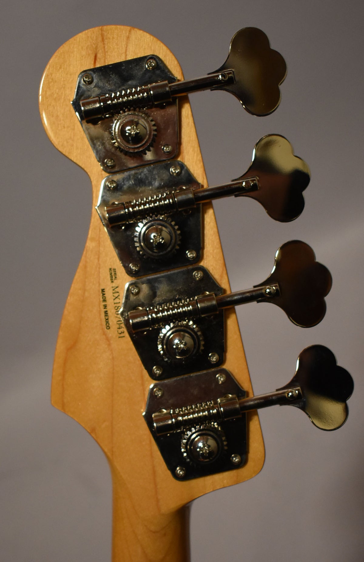 Close-up of the bass headstock's rear, showing four chrome clover-leaf tuners on a light wood finish, with visible serial number.