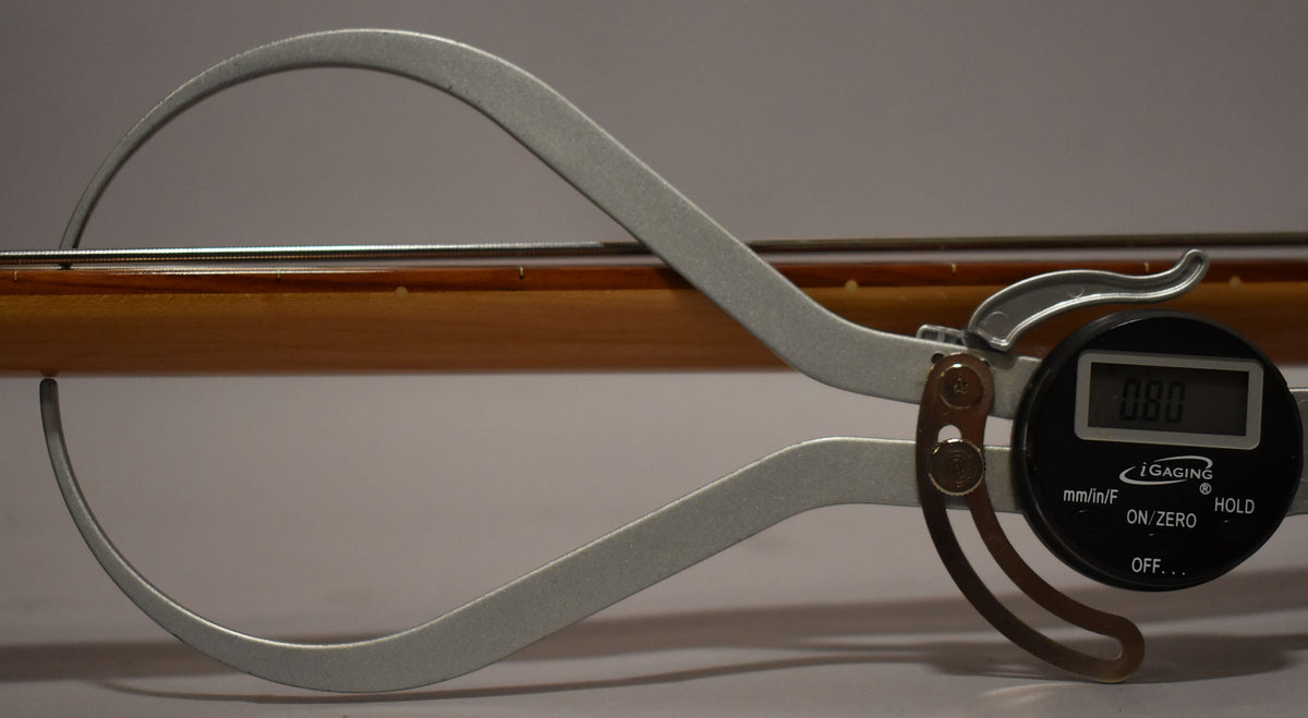 A digital caliper measures the fretless bass neck's relief, showing 0.00 on the display.