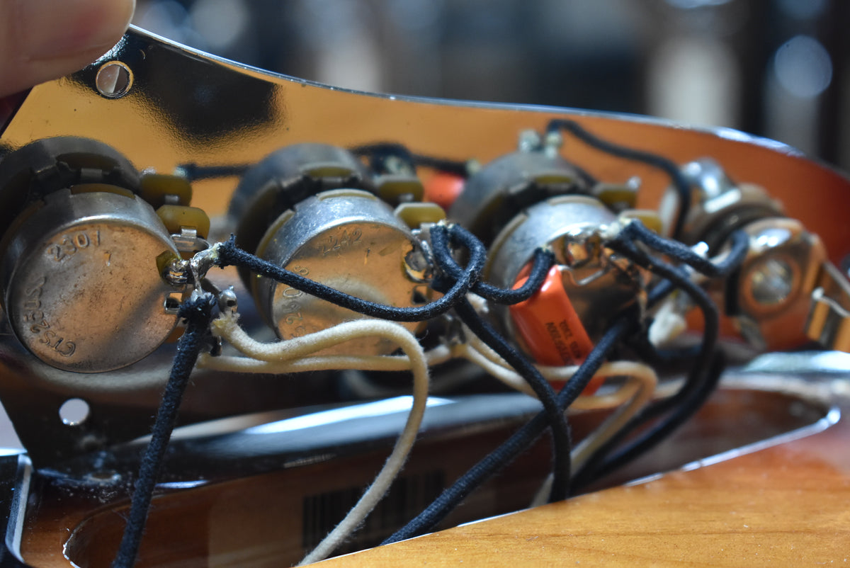 Close-up of the bass's control cavity, revealing three silver pots, orange capacitors, and cloth-wrapped wiring on a chrome plate.