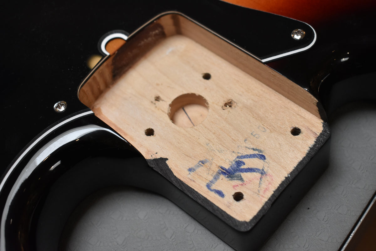 Close-up of the bass's neck pocket, showing the bare wood with screw holes and blue markings, against the glossy sunburst body.