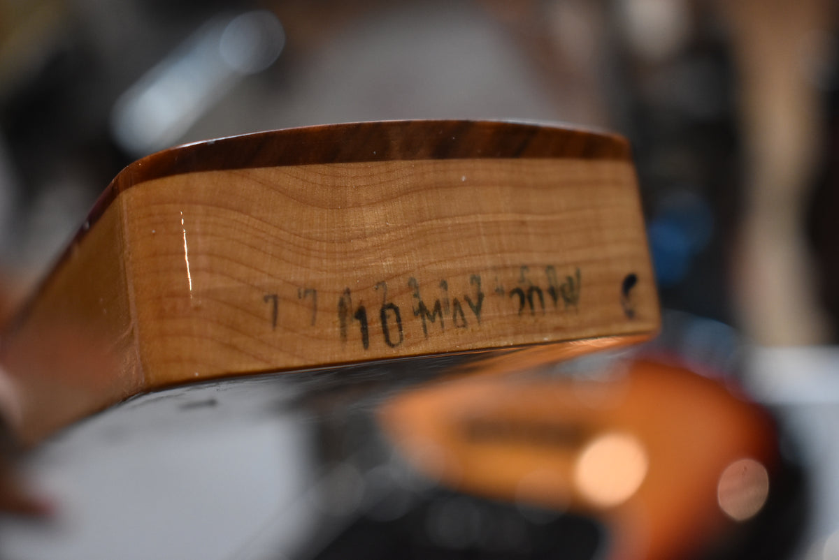 Close-up of the light wood neck heel, showing "10 MAY 2018" stamped in black ink, with a sunburst body blurred in the background.
