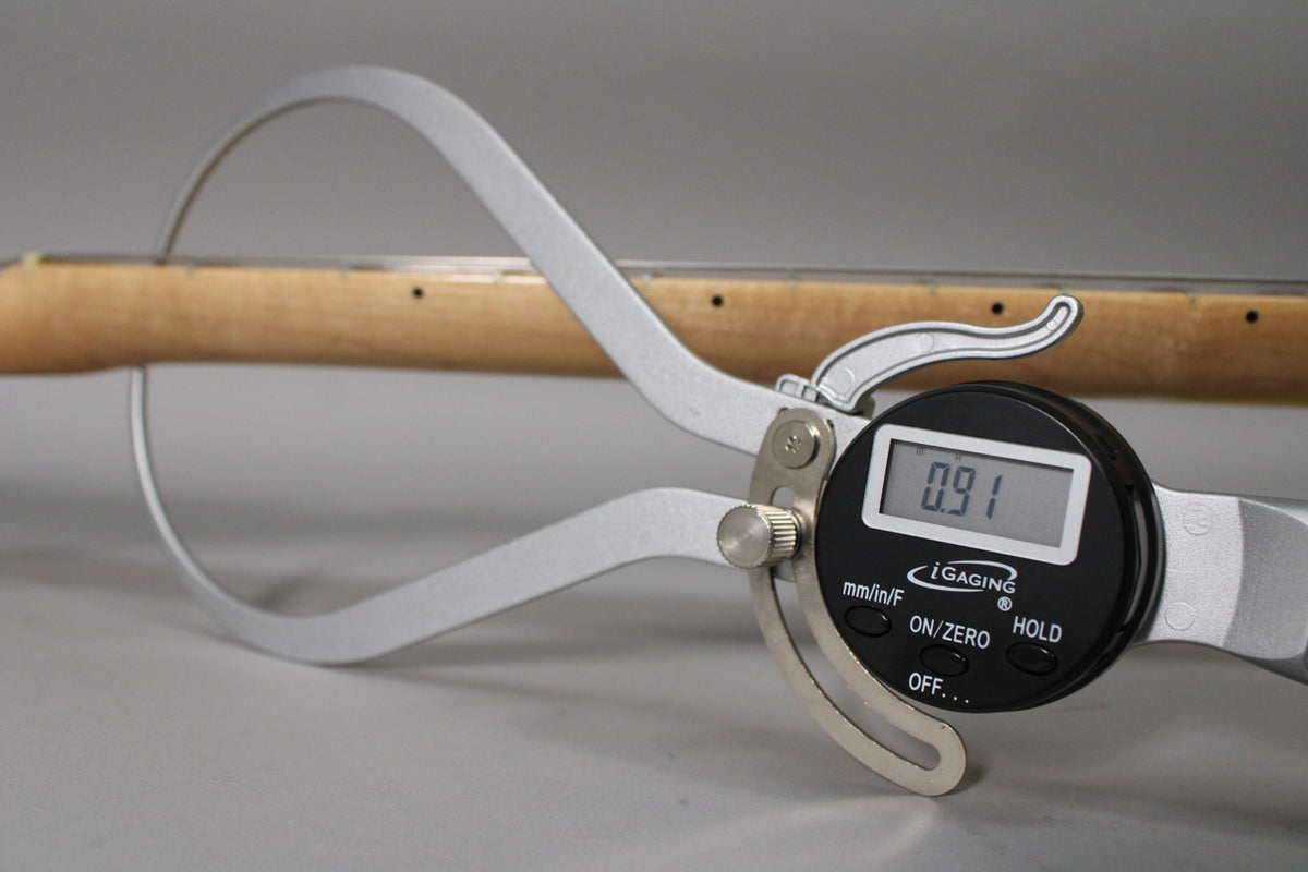 A digital caliper measures the guitar neck, showing a reading of 0.91, with the light wood neck visible behind it.