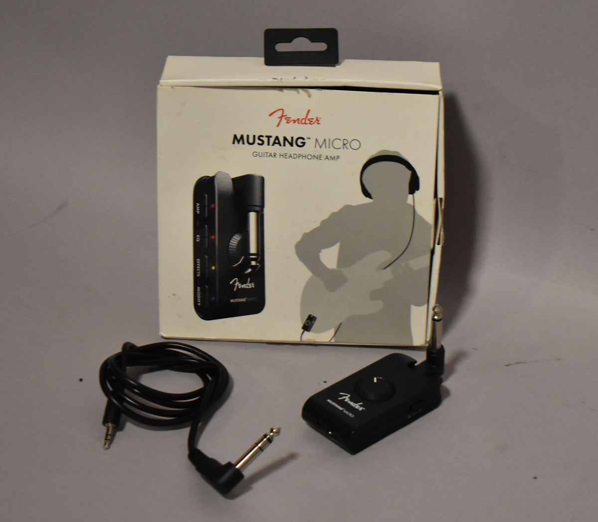 The black headphone amp, coiled cable, and box are angled on a grey surface, showing the amp's input jack.