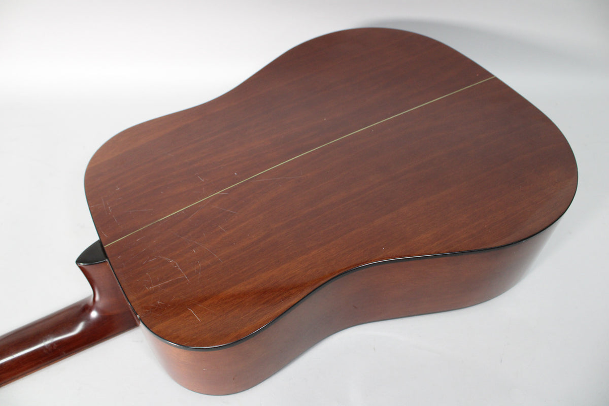 The acoustic guitar's dark brown back and sides are visible, with a center seam and many surface scratches and scuffs.