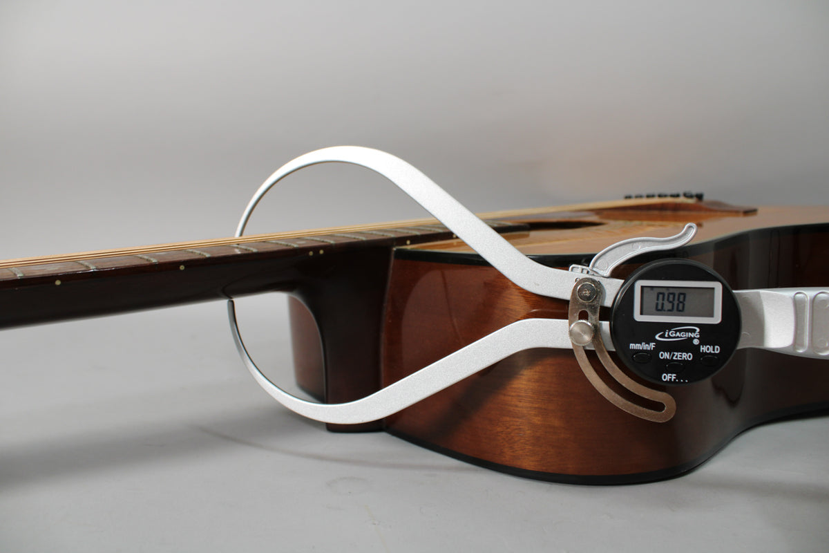 A digital caliper measures the dark brown neck joint of the natural wood finish acoustic guitar, displaying "0.98".