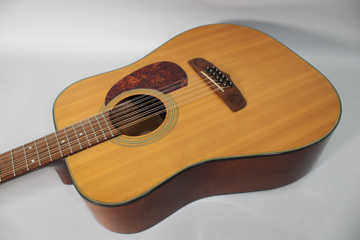 The 12-string acoustic guitar's light-colored top, dark sides, and tortoiseshell pickguard are visible from a high angle.