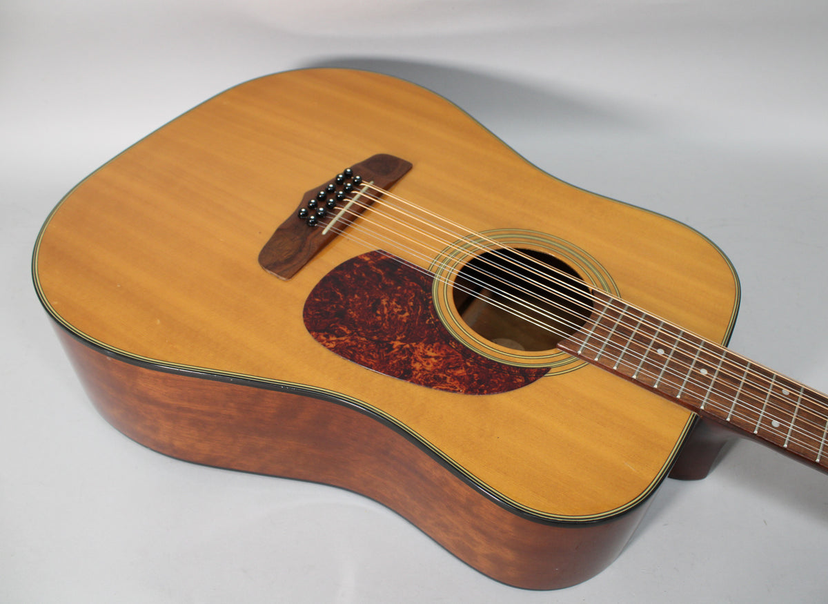 A light brown 12-string acoustic guitar, angled from the upper left, showing the body, pickguard, bridge, and part of the fretboard.