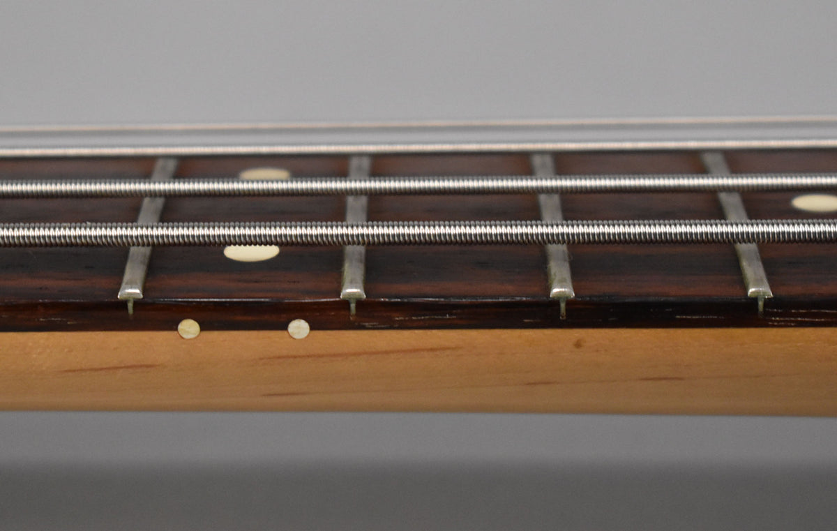 Close-up of the bass neck, showing the dark fretboard with oval inlays, four strings, and circular side dots on the light wood.