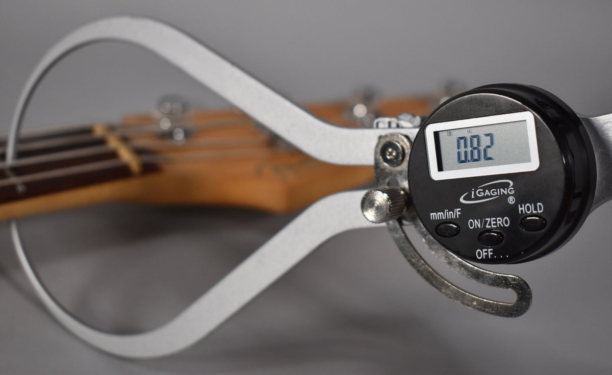 A digital caliper measures the bass's neck thickness at 0.82 inches, with the headstock and tuners blurred in the background.