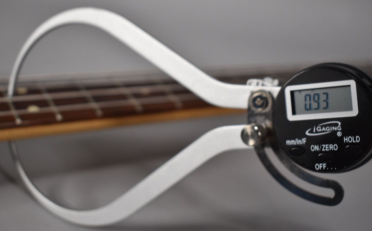 A digital caliper measures the bass's fretboard, displaying "0.93" on its screen, with visible fret wear.