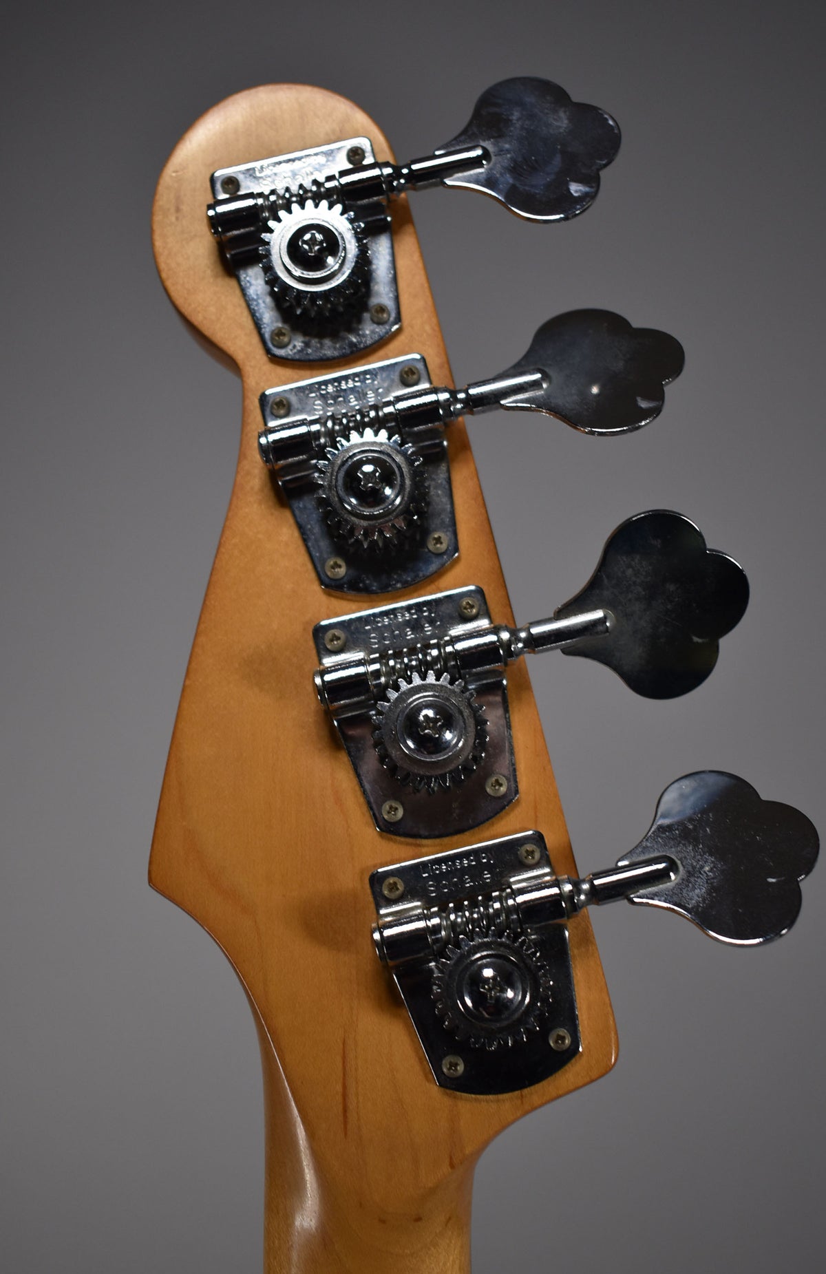 Close-up of the bass headstock from behind, showing four chrome clover-leaf tuners on a light wood finish, with minor scratches.