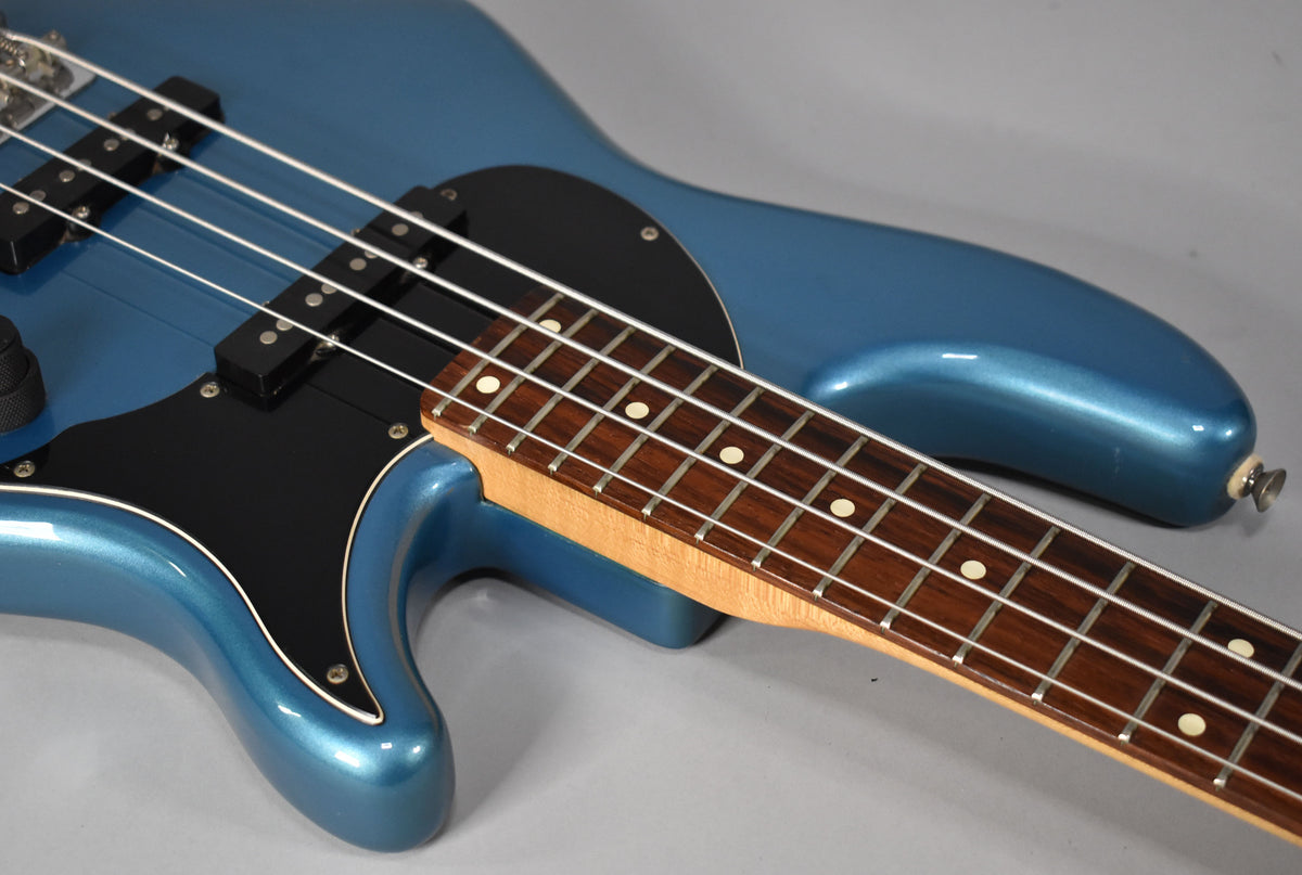 Close-up of a metallic blue bass body with a black pickguard, two pickups, and a rosewood fretboard with dot inlays.