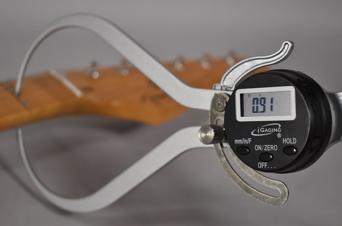 A digital caliper measures the neck thickness at the first fret, showing 0.91, with the headstock blurred in the background.