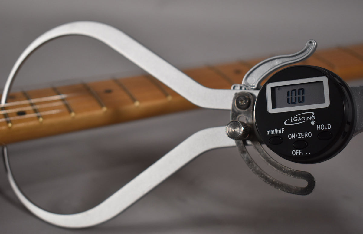 A digital caliper measures the guitar's maple fretboard, showing "1.00" on its screen, with frets and strings visible.
