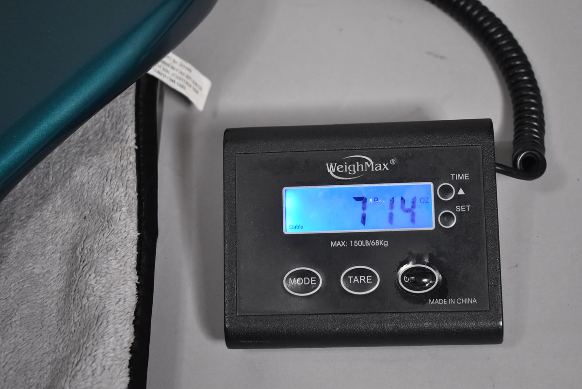 The guitar's Ocean Turquoise body rests on a gray gig bag, with a digital scale displaying "7 lbs 14 oz" in the foreground.