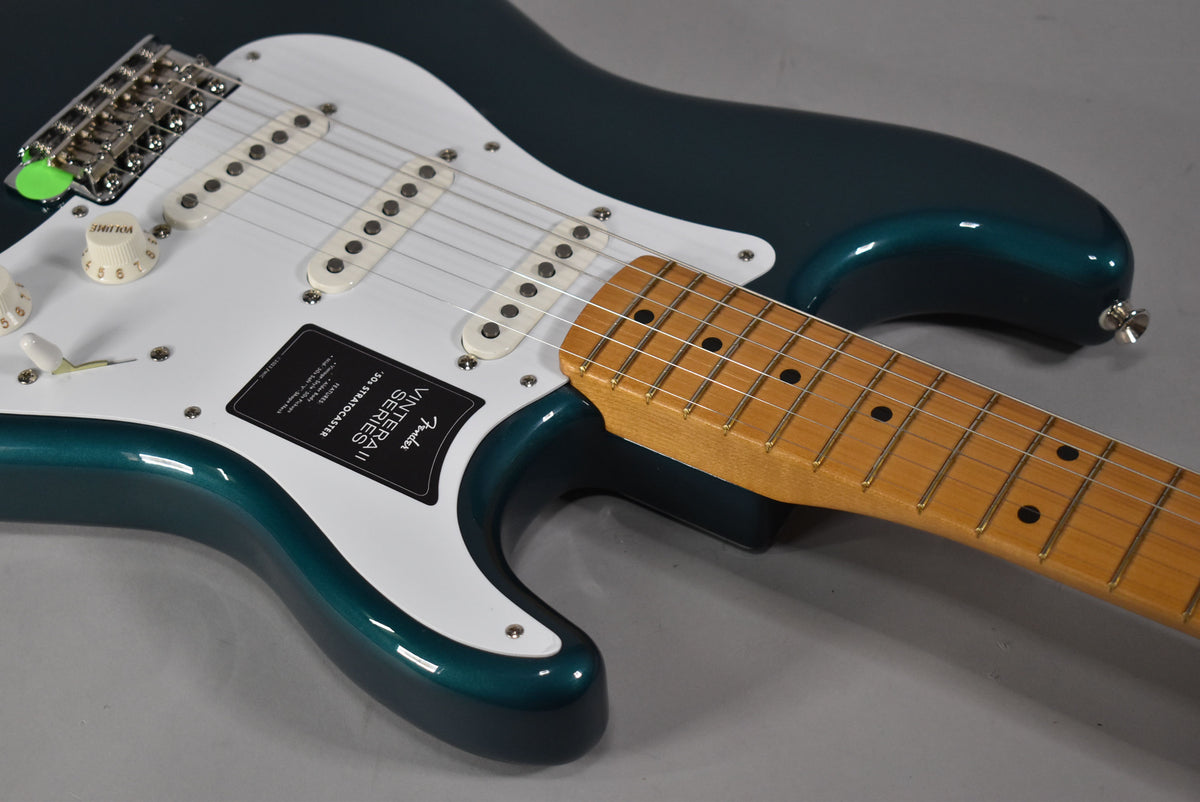 The glossy ocean turquoise guitar body with a white pickguard and three single-coil pickups is angled, showing the maple fretboard.