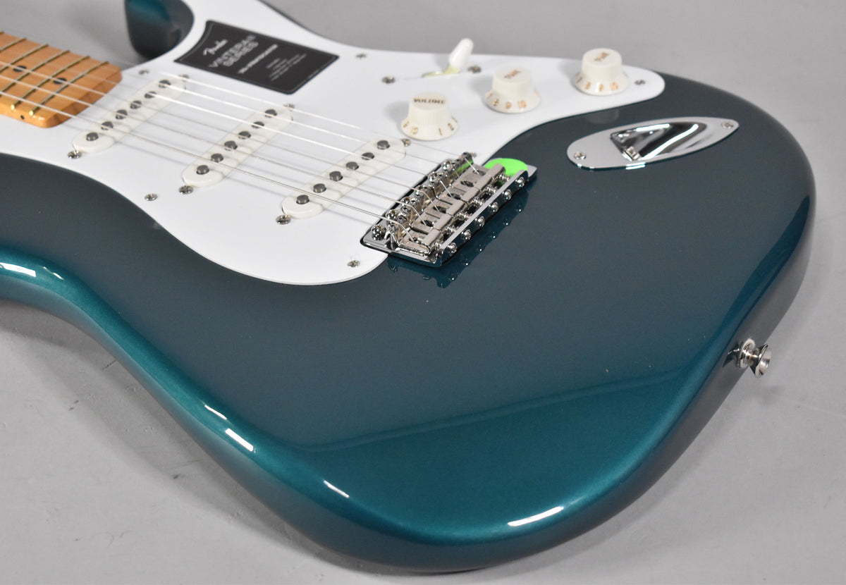 The guitar's glossy ocean turquoise body is angled from the lower right, revealing a white pickguard, three single-coil pickups, and cream-colored control knobs.
