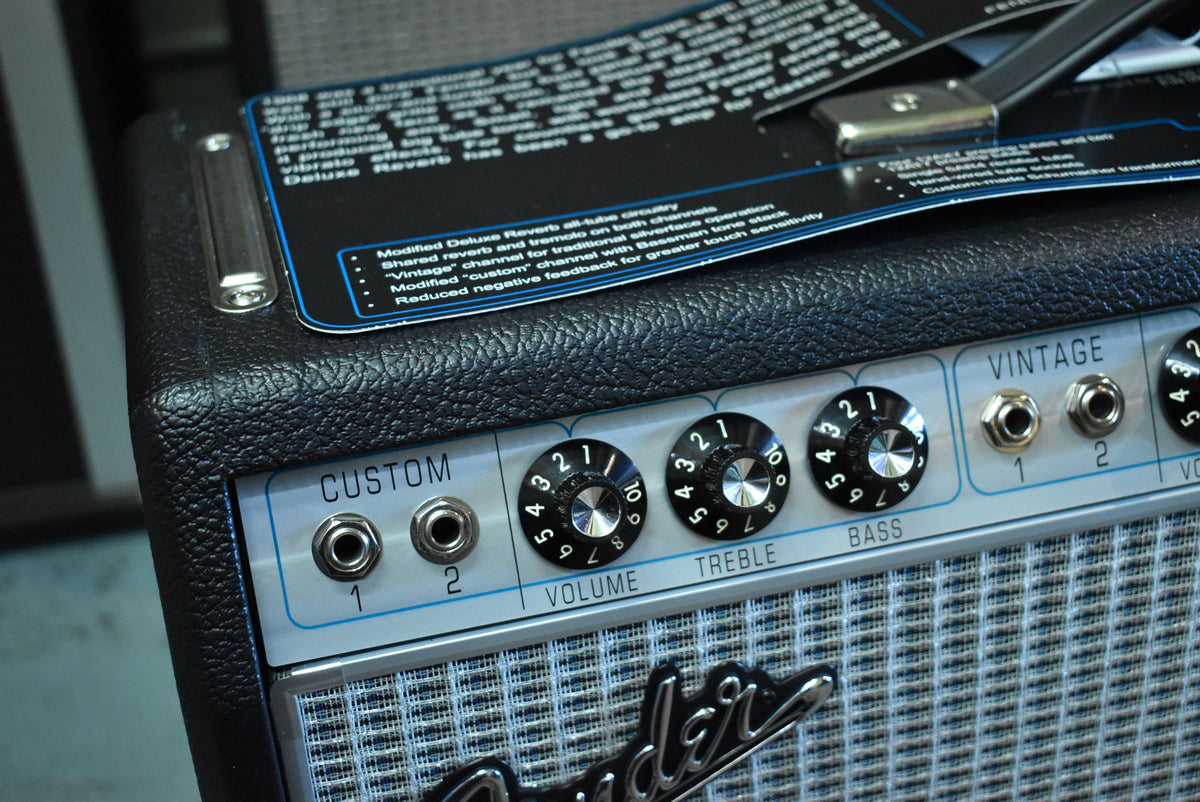 Close-up of the black textured amp's silver control panel, showing input jacks, black numbered knobs, and a hang tag on top.