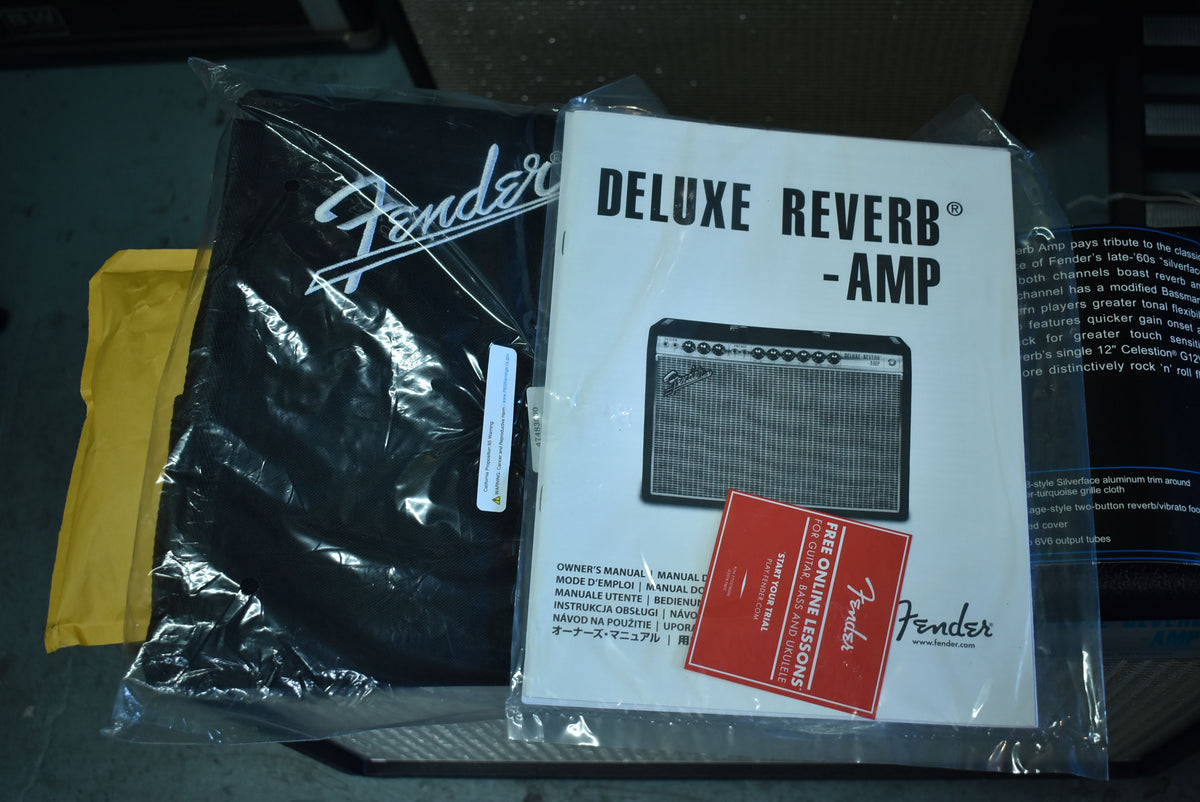 A black amp cover and owner's manual, both sealed in plastic, are shown from above, with a yellow envelope to the left.