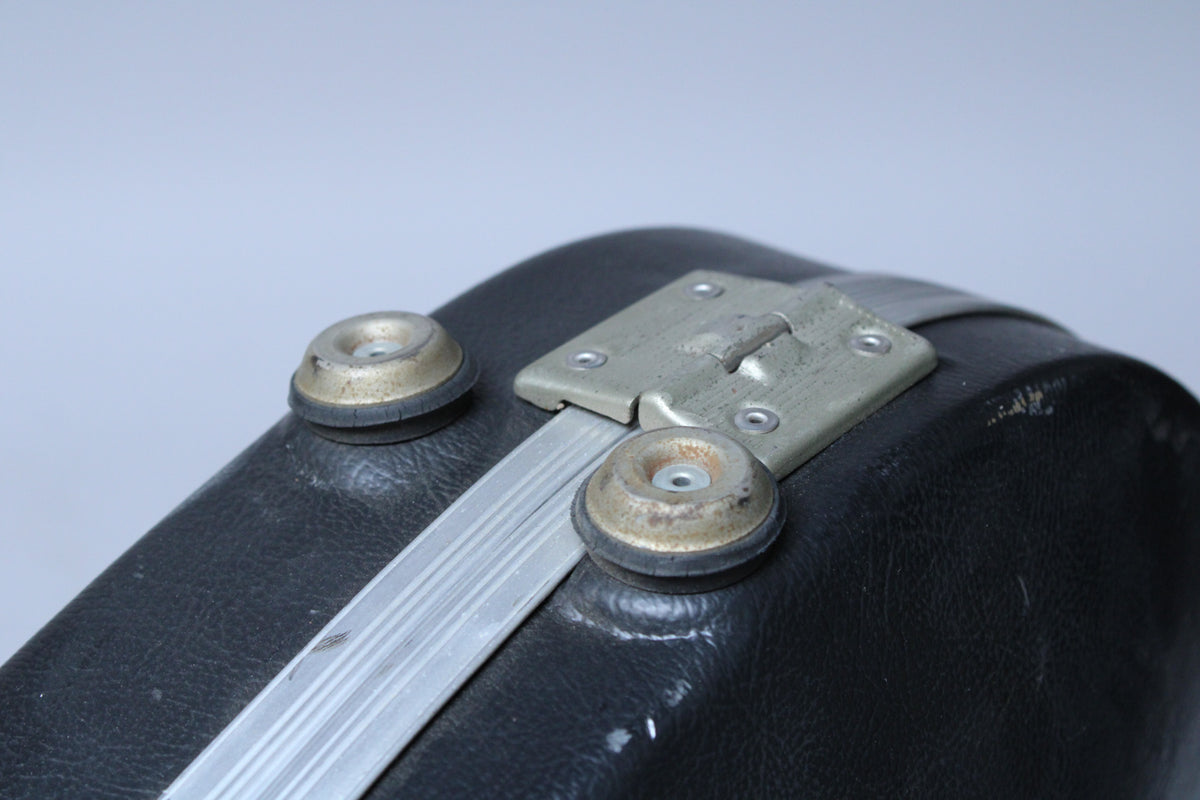 Close-up of the black case's textured surface, showing two worn metal feet, a silver-toned latch, and a long aluminum strip.