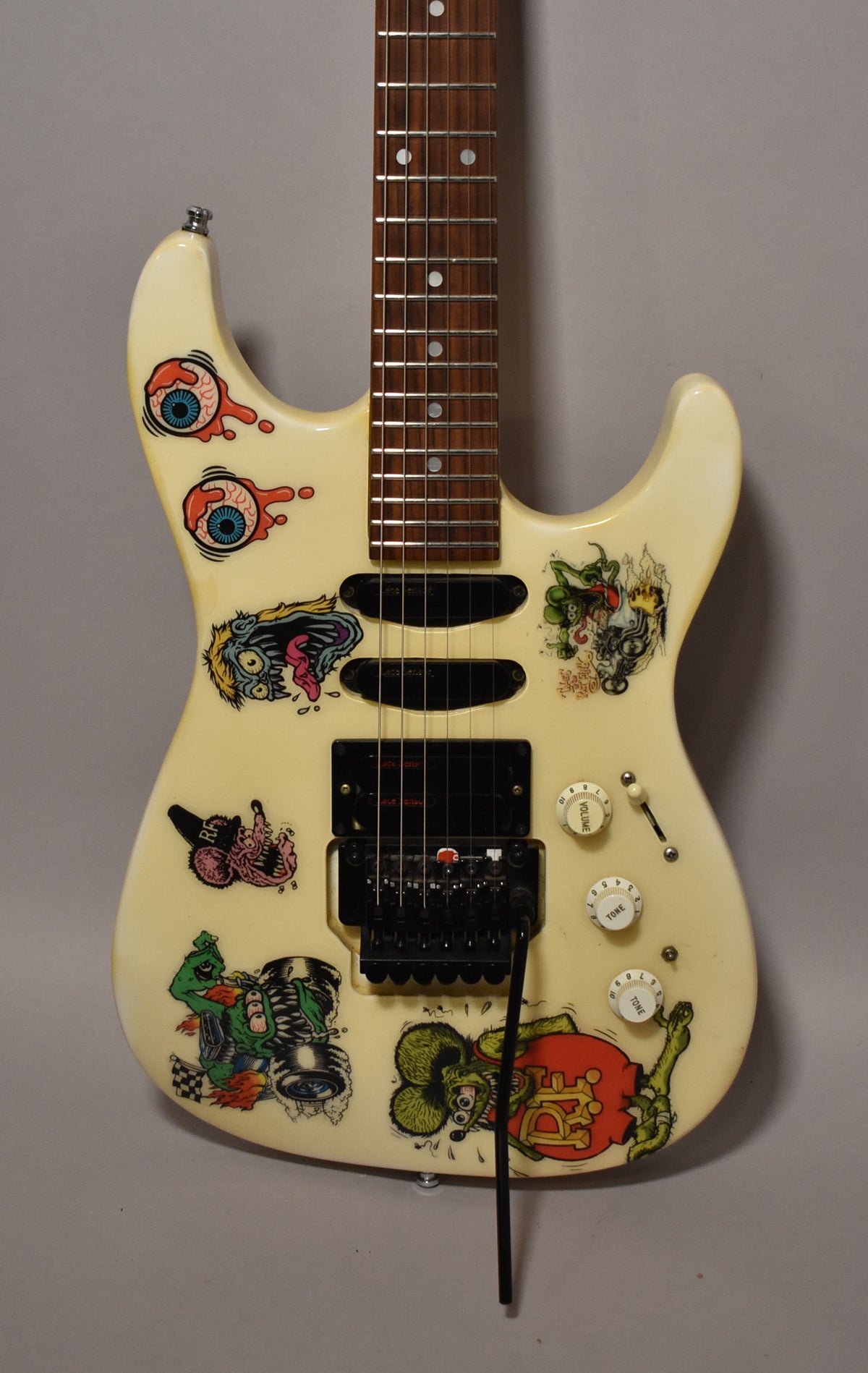 The cream-colored electric guitar body, covered in colorful monster stickers, is angled slightly, revealing three black pickups and a tremolo arm.