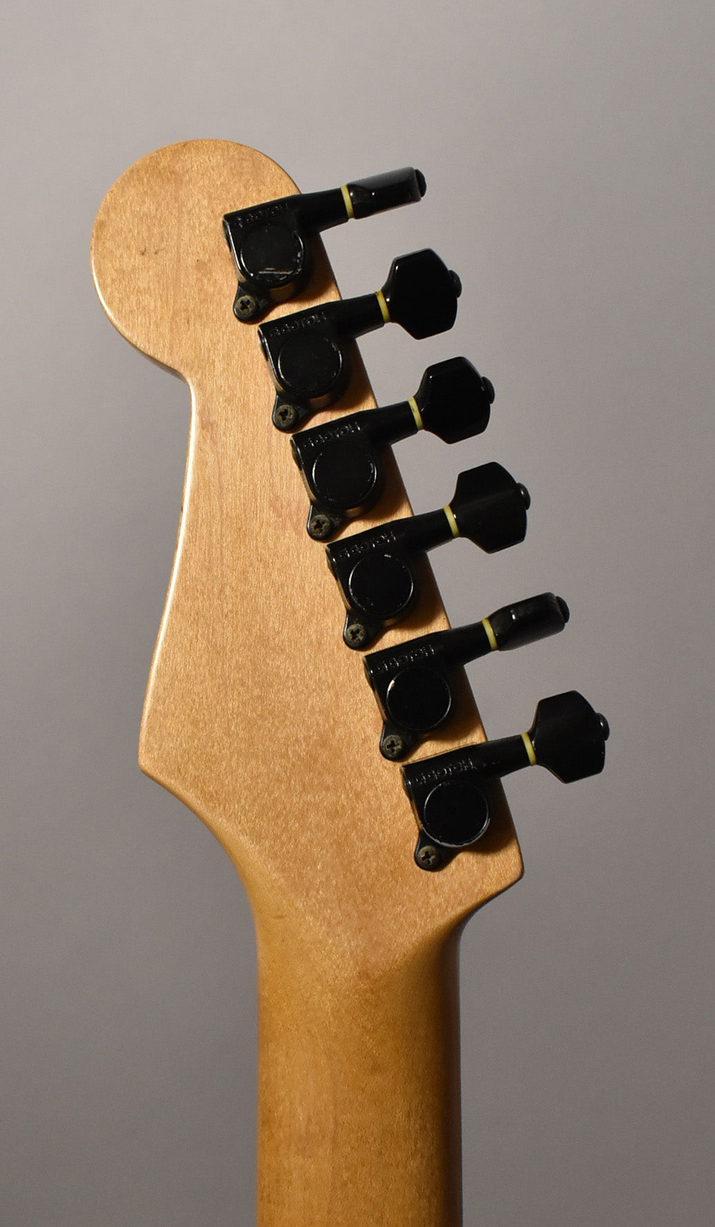 The guitar's light wood headstock and neck are seen from behind, with six black tuning machines showing some wear.