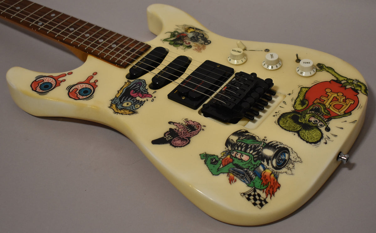 Angled close-up of a white electric guitar body covered in colorful monster stickers, with three black Lace Sensor pickups and white control knobs.