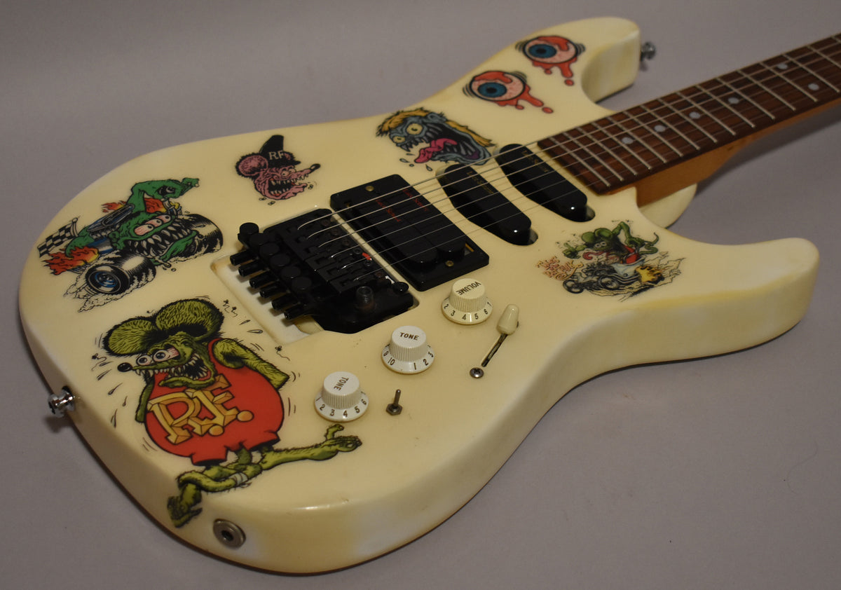 The white electric guitar's body is covered in colorful Rat Fink and monster car stickers, with black Lace Sensor pickups and a Floyd Rose tremolo.