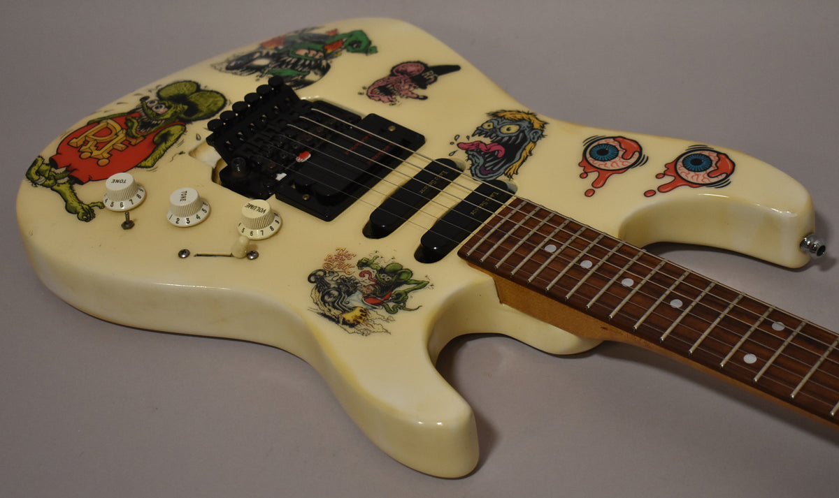 The white electric guitar's body, covered in colorful monster stickers, is angled to show its black pickups, tremolo, and three white control knobs.