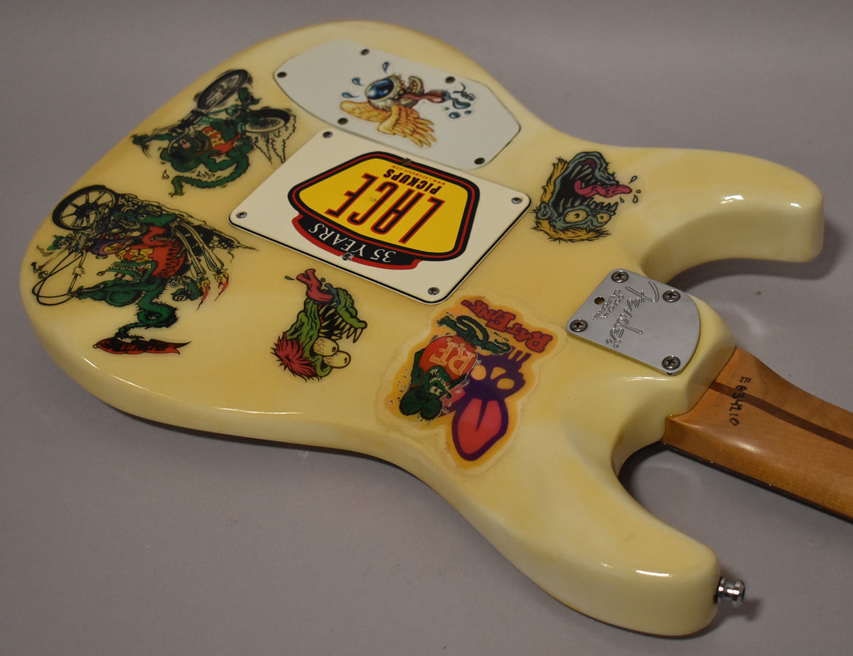 Rear view of a white electric guitar body covered in colorful cartoon stickers, with a chrome neck plate and output jack.