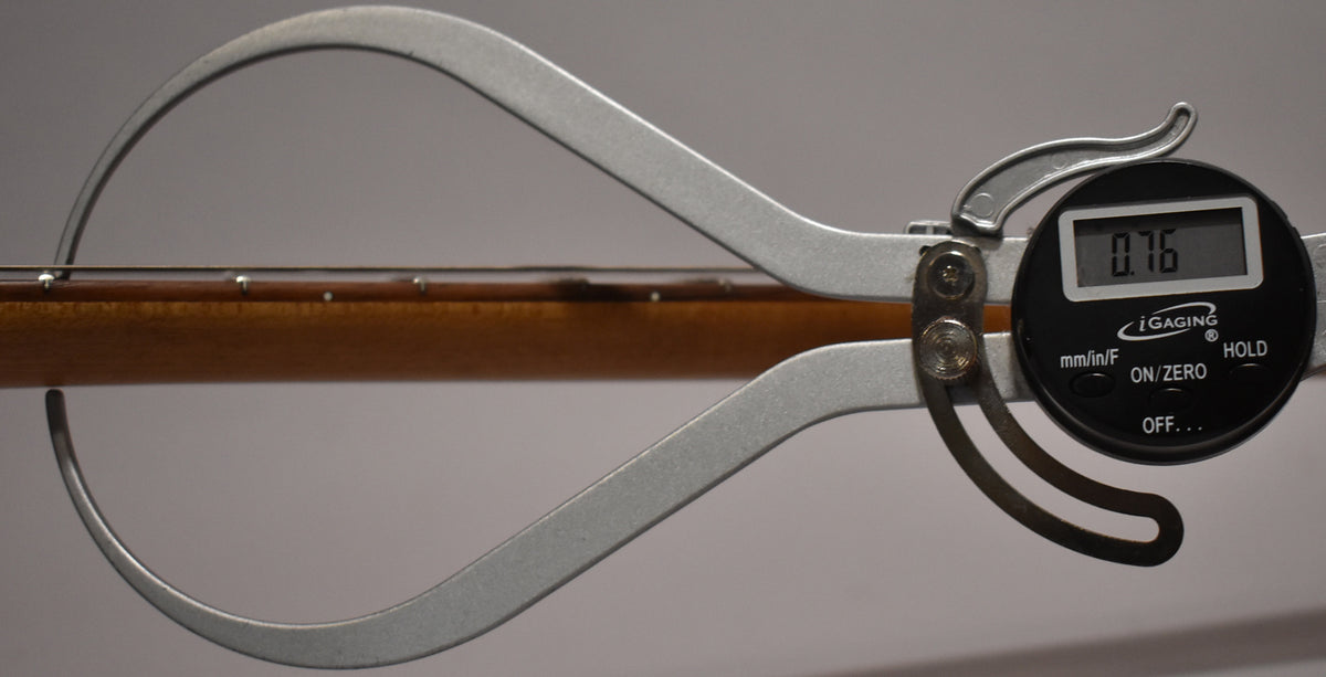 A digital caliper measures the guitar's neck, showing 0.76 on its screen, with the fretboard and strings visible.