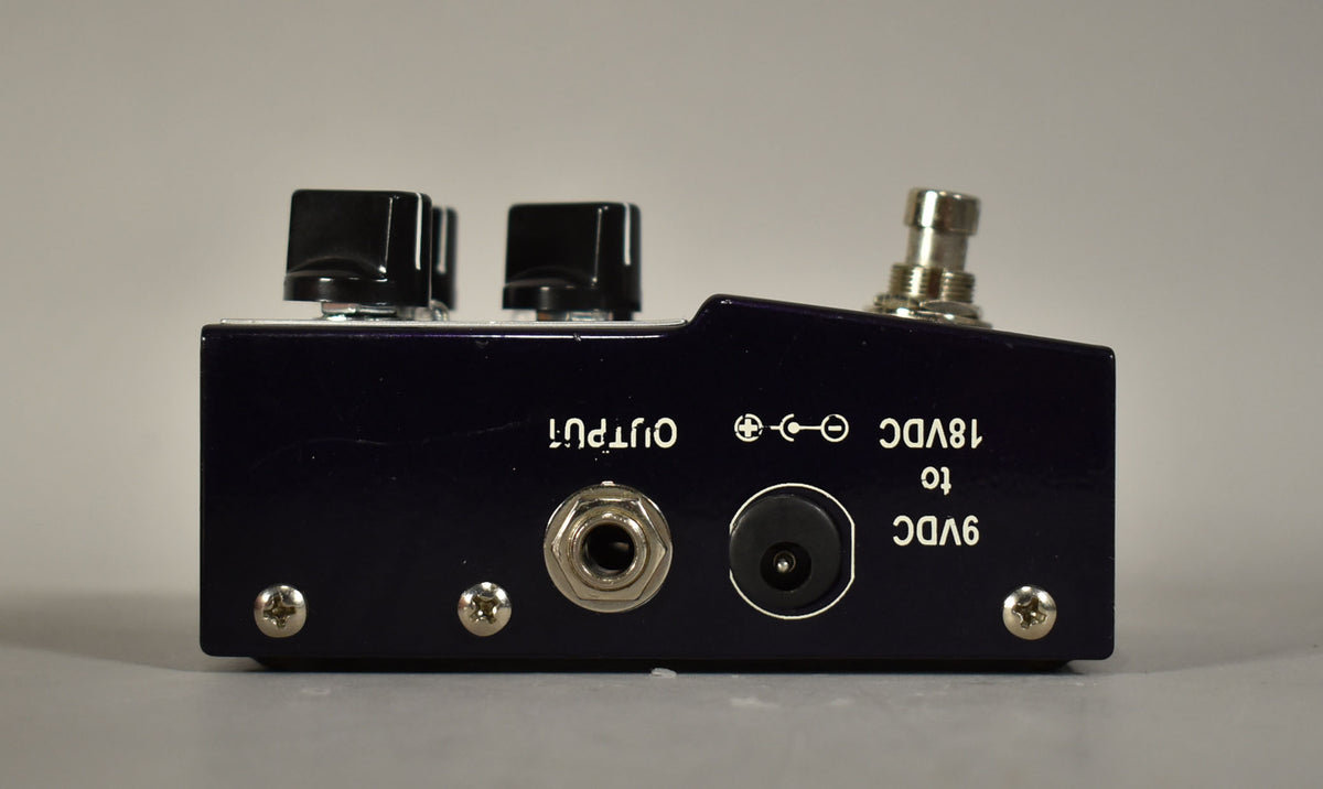 The pedal's dark purple side panel, angled slightly, shows an output jack, power input, and two black control knobs.