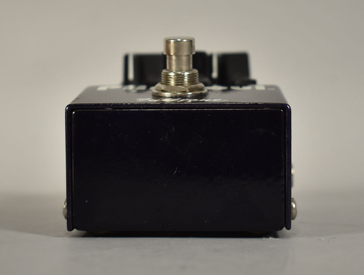 The dark purple pedal is viewed from the front, showing the silver footswitch and black control knobs on top.