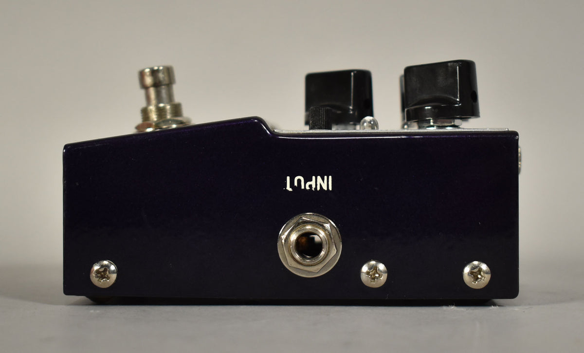 The dark purple pedal is viewed from the side, showing the input jack, two black knobs, and a silver footswitch.
