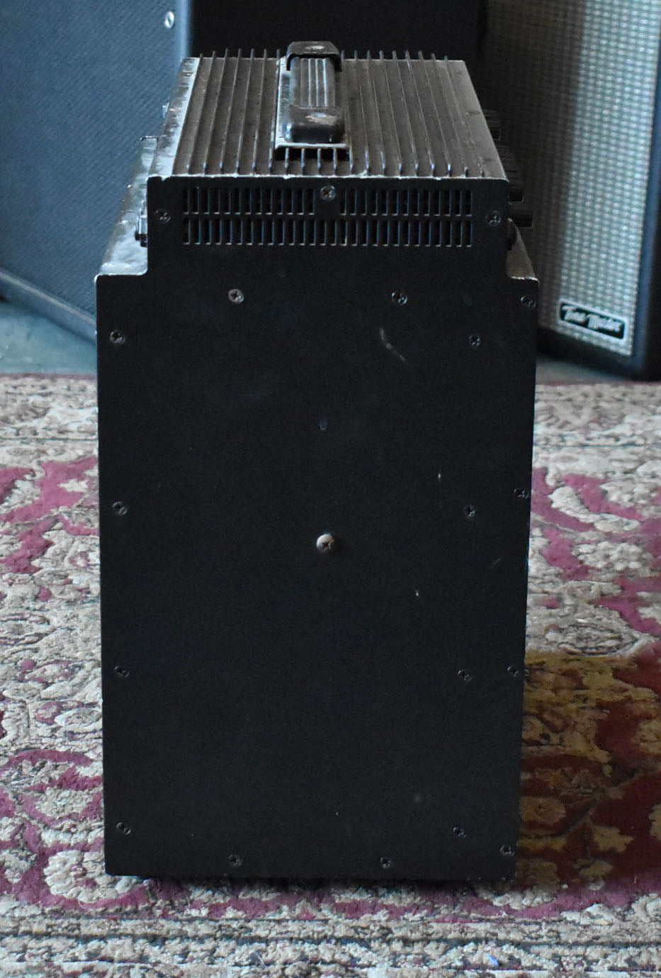 The black amplifier is viewed from the side, showing a textured finish, exposed screws, and a top cooling fin array with a handle.