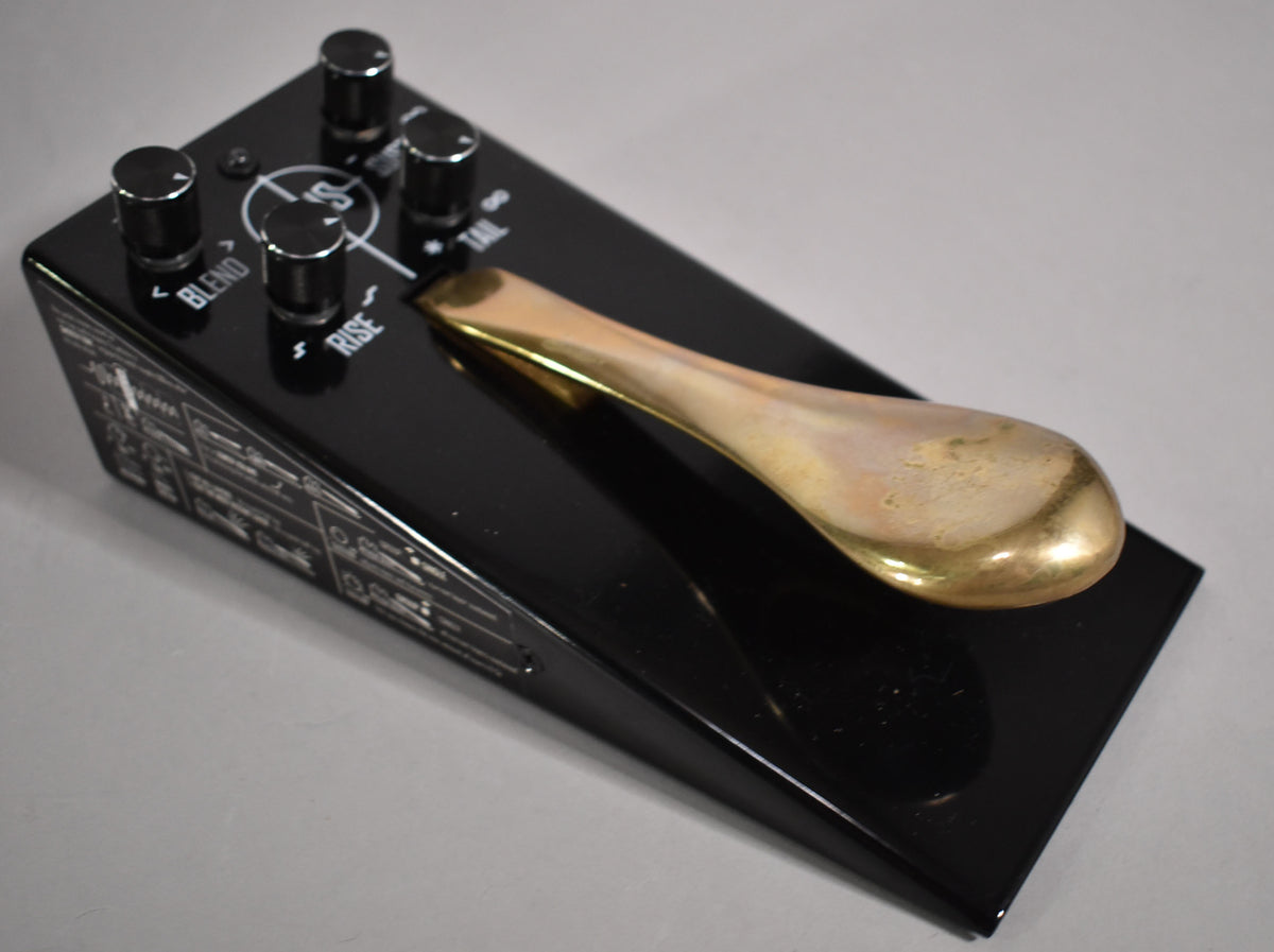 A black pedal with a glossy finish, angled from the left, showing four silver knobs and a tarnished brass-colored sustain pedal.