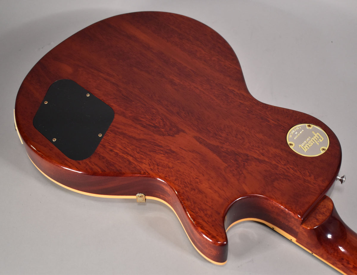 The back of the guitar shows a rich reddish-brown wood grain, a black control cavity cover, and a gold medallion.