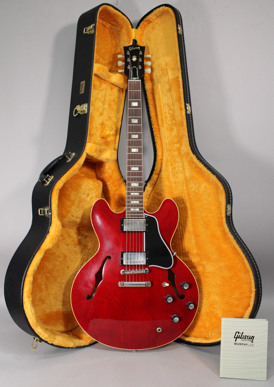 Full-length view of a cherry red semi-hollowbody electric guitar with f-holes, chrome hardware, and a black pickguard, standing in its open case.