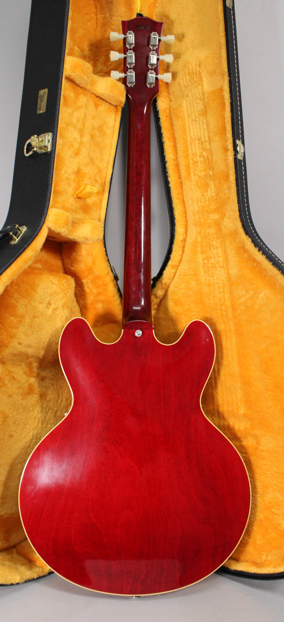 The back of a cherry red semi-hollow guitar with a light aged finish, showing the neck, headstock with six tuners, and open yellow-lined case.