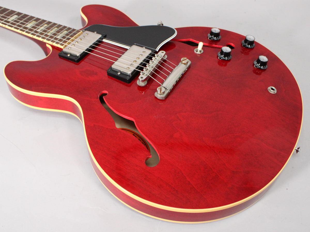 The cherry red semi-hollow guitar is angled from the upper left, showing its body, F-hole, pickups, and four black control knobs.